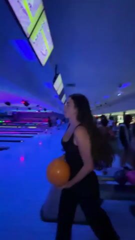 A woman in a black dress is holding an orange bowling ball in a bowling alley. The bowling alley is dimly lit with blue and purple lights. The woman turns her head to her left and smiles. She then turns to her right and begins to walk away from the camera, holding the bowling ball. The bowling lanes are visible in the background.