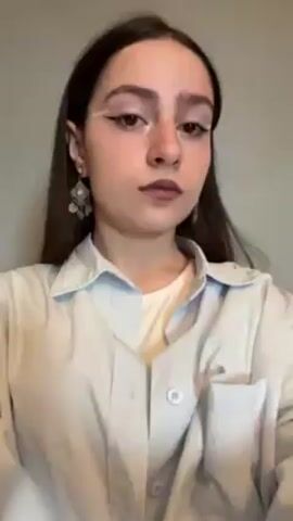 A young woman with long, dark hair and distinctive white eyeliner stands indoors against a neutral background. She wears a light blue, buttoned-down shirt over a white t-shirt, and dangle earrings. She poses for the camera, moving her hand near her face, showing off a ring.