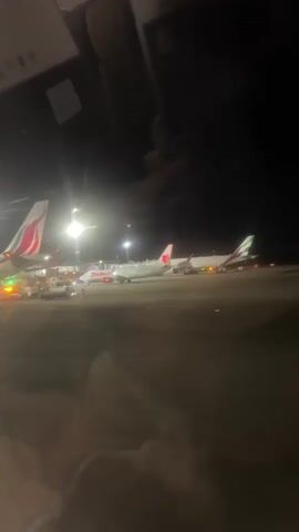 The video shows an airport tarmac at night. Several airplanes are parked on the ground, illuminated by floodlights. The camera appears to be inside another aircraft or a terminal, looking out a window. The lighting is dim, and the overall scene is dark with some reflections on the glass.