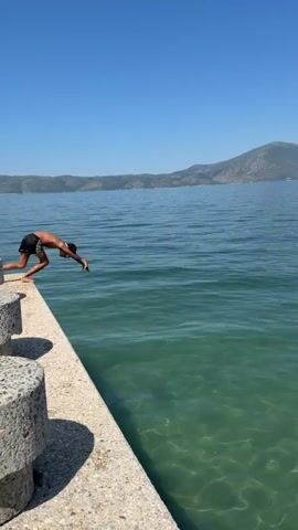 A person wearing black swim trunks jumps into clear, blue-green water from a concrete pier. The person is captured mid-air, facing away from the camera, with arms outstretched. Ripples and a splash are visible on the water's surface as the person enters the water. The background shows a calm body of water and a distant, hilly landscape under a clear blue sky.