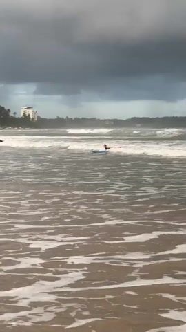 The video shows a group of people learning to surf in Weligama, Sri Lanka. The scene is set on a beach with choppy waves, suggesting overcast or stormy weather indicated by the dark clouds in the sky. Several individuals are in the water, some on surfboards and others paddling. One person, wearing a black and yellow wetsuit, is attempting to stand on a blue surfboard. Another individual, in a black wetsuit, is further back in the water with a surfboard. In the background, a white building, likely a hotel, is visible behind a line of palm trees. Several fishing boats are also pulled up on the shore. The overall mood appears to be focused on the activity of surfing, with no overt displays of emotion. The time of day seems to be daytime, given the light, though the cloud cover creates a subdued atmosphere.