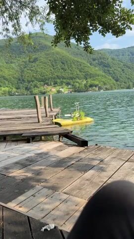 The media file shows a serene lake scene on a sunny day. In the foreground, a rustic wooden dock juts out into the shimmering green water. Several wooden posts rise from the dock, suggesting it's a well-used landing for boats. A bright yellow object, possibly a small pedal boat or a floating platform, is moored to the dock and is partially visible in the water. It has some greenery growing on it and a metal pole with a small antenna-like structure.

The lake itself is calm, with gentle ripples on its surface. Beyond the lake, rolling green hills covered in dense forest rise steeply. Nestled within the hills, small houses with red roofs are visible, hinting at a peaceful, rural community. The overall impression is one of tranquility and natural beauty. The time of day appears to be daytime, with bright sunlight casting shadows on the dock, and the weather seems pleasant. The location cues, along with the style of the houses and the surrounding landscape, strongly suggest a setting in Bosnia and Herzegovina, specifically Ćusine. No people are visible in the frame, adding to the sense of quiet solitude. The overall mood is peaceful and inviting, evoking a sense of summer relaxation. There is no visible text in the media file.