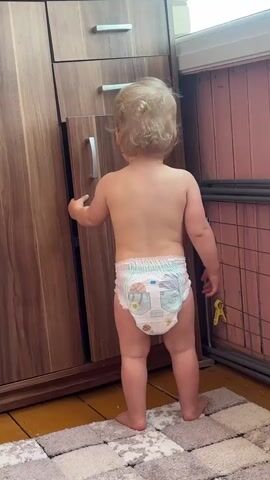 A toddler wearing a diaper stands facing away from the camera, their back to the viewer. They are reaching for a partially open cabinet door. The cabinet is made of wood and has a silver handle. The toddler's hair is blonde and curly. The floor is covered with a rug with a checkered pattern. To the right of the cabinet, a metal railing or shelf is visible, with a yellow clothespin attached to it. The background appears to be an indoor setting, possibly a room.