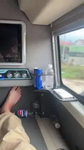 Inside what appears to be a bus, a person is lying down. A bare foot is visible. There's a small television screen, a Pepsi bottle, and a can of soda in a cup holder nearby. A hand walks its fingers across the bus window while looking out at the scenery passing by. Buildings and greenery are visible outside.