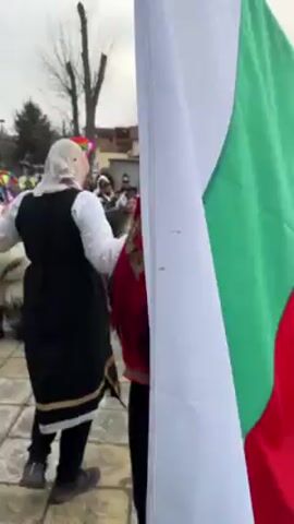 A group of people is dancing in a public square. They are dressed in folk costumes, with white shirts, dark vests, and patterned skirts or pants. Some wear head coverings, including scarves, hats, and elaborate headdresses. A few individuals wear large, shaggy costumes that obscure their faces. A Bulgarian flag is held aloft among the dancers. The square is paved with stone and is bordered by buildings. Other people, some wearing masks, are present in the background. The sky is overcast.