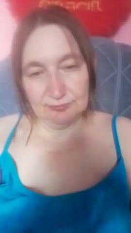 A medium shot shows a middle-aged Caucasian woman with brown hair, wearing a blue tank top. She is seated, facing the camera, and appears to be in a room with a pink wall and a red heart-shaped pillow in the background. The video is slightly blurry.