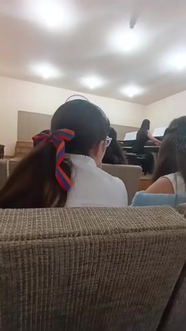 The video shows a room with several people. In the foreground, two people are seated on couches, facing away from the camera. One person has long, dark hair tied in a ponytail with a red, orange, and blue ribbon. In the background, a person is playing a piano. Another person is seated at a second piano, looking at sheet music. The room appears to be a concert hall or a performance space, with tiered seating visible on the left side. The lighting suggests an indoor environment, likely during the day or with artificial lighting.