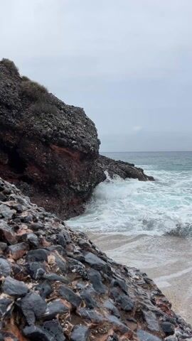 The video shows waves crashing against rocks on a beach. The sky is overcast and grey, suggesting it is a cloudy day. The sea is a turquoise-blue color with white foam on the waves. The rocks are dark grey, black, and red. Some vegetation can be seen growing on the cliff. The camera pans slightly to the right to show the ocean.