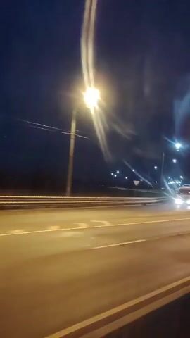 The video shows a nighttime view from a vehicle moving along a highway. Streetlights illuminate the dark sky and the road ahead. The blur of motion suggests the vehicle is traveling at a considerable speed. The road has multiple lanes, separated by white lines, and a median barrier is visible. The presence of streetlights and the lack of natural light indicate it is nighttime or very early morning. In one frame, a car with illuminated headlights is seen passing by.