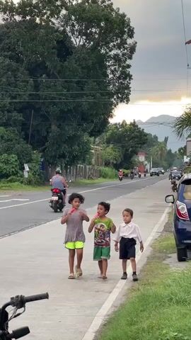 The video shows three children walking on a sidewalk next to a road. The child on the left is wearing a pink shirt and yellow shorts and holding a red popsicle. The child in the middle is wearing a green shirt and green shorts and holding a red popsicle. The child on the right is wearing a white shirt and black shorts and black shoes. There are trees and buildings on the left side of the road, and more trees and mountains in the distance. There are several motorcycles and a car on the road. The sky is cloudy.
