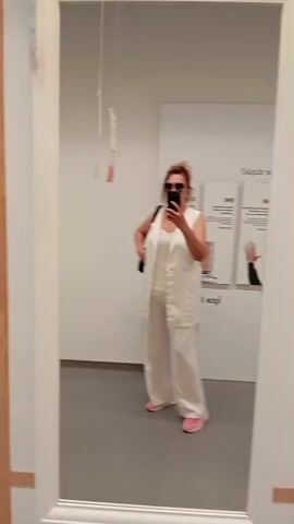 The video begins with a woman in a white outfit and sunglasses taking a selfie in a large mirror. She is standing in what appears to be an IKEA store, as indicated by the IKEA logo visible in the reflection and on a tag hanging from the ceiling. The woman is wearing a white sleeveless vest over a white top and white wide-leg pants, with pink slippers. The reflection shows two price tags hanging from the ceiling. The camera then pans to the left, revealing a wall with a wooden grid design. Two framed pictures with white mats and black frames are mounted on the wall. The camera continues to pan left, showing more framed pictures and decorative baskets mounted on the wall, as well as a circular wooden mirror. The aisle appears to be an exhibition space within the store, with a gray floor and white walls.