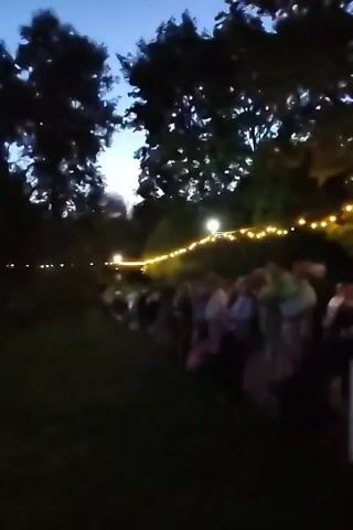 The video shows a dimly lit outdoor scene at dusk or night. A line of people is visible, silhouetted against the dark background of trees. String lights, emitting a warm yellow glow, are draped along the top of a fence or railing, creating a festive atmosphere. The lights are also reflected on the surface of what appears to be a body of water in the foreground, adding to the ambiance. The sky is a deep blue, indicating the transition from day to night. The overall impression is of a gathering or event taking place in a natural setting, illuminated by decorative lighting.