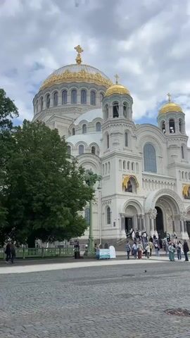 The video shows the exterior of a large, ornate white cathedral with golden domes under a cloudy sky. The cathedral has a grand entrance with steps leading up to it, and several smaller golden domes on towers. A large green tree is visible to the left of the cathedral. Several people are walking on the cobblestone plaza in front of the cathedral and ascending the steps. There is a lamppost in the foreground and some signs. The scene is static with no discernible movement.