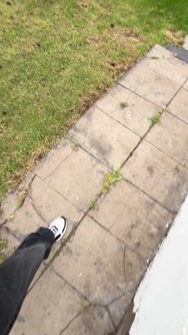 A young man is walking on a sidewalk in a suburban neighborhood while filming himself with a handheld device. He's wearing a black hoodie, dark jeans, and white sneakers. He also has a brown crossbody bag with a repeating pattern. The sidewalk is adjacent to a grassy lawn, and across the street are houses. The sky appears overcast. The camera angle is slightly downward, capturing the man's face and upper body, as well as his legs and the sidewalk in front of him. He seems to be walking at a moderate pace.