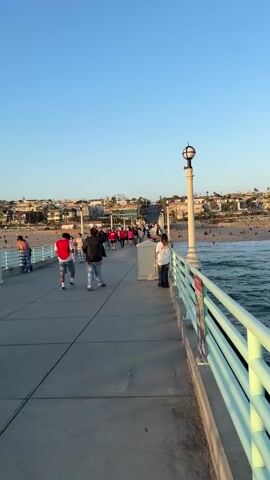 A vertical video captures a busy pier on a sunny day. A concrete walkway stretches into the distance, lined with a light green railing. People are walking in both directions on the pier. On the right side of the pier, the ocean with moderate waves is visible. On the left side of the pier, a sandy beach with scattered people and buildings can be seen in the background. The sky is clear and blue. A lamppost with a decorative top stands prominently on the right side of the pier. Several individuals are captured from behind as they walk away from the camera, including a person wearing a red vest and another in a white shirt. In the foreground, two people are standing near the railing, facing away from the camera, observing the scene. Another individual wearing a black t-shirt with white lettering and gray and white patterned pants stands to the left, looking towards the pier. The overall atmosphere suggests a lively recreational area.
