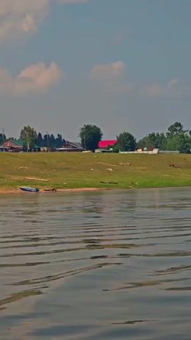 The video shows a body of water with ripples on the surface. On the shore in the distance, there are several buildings and trees. On the grassy bank, there is a small green boat, hay bales, and a person standing. Further down the bank, there are some vehicles, including a truck towing a trailer and a white van. The sky is partly cloudy.