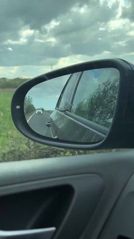 From inside a car, a side mirror reflects the traffic behind. The road is bordered by green vegetation and trees on either side. The sky is cloudy. The video is shaky and not very clear.