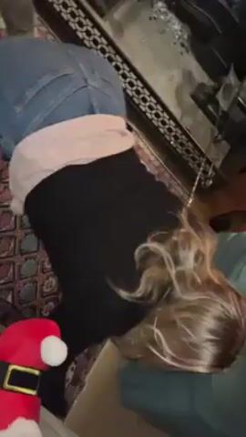 A person wearing jeans, a pink top, and black leggings is on their hands and knees on a patterned rug. The rug has various colors and geometric shapes. Next to the person is a Santa Claus doll. Behind the person is a fireplace with a black metal grate. There are scattered pieces of white material near the fireplace. The person appears to be reaching towards something near the fireplace.