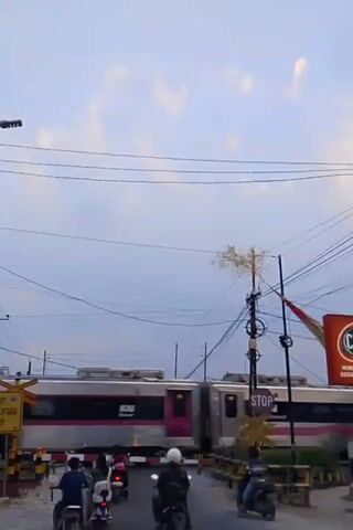 A train passes through a railroad crossing. Several people on motorcycles are stopped at the crossing, waiting for the train to pass. The sky is overcast, and there are power lines strung across the scene.