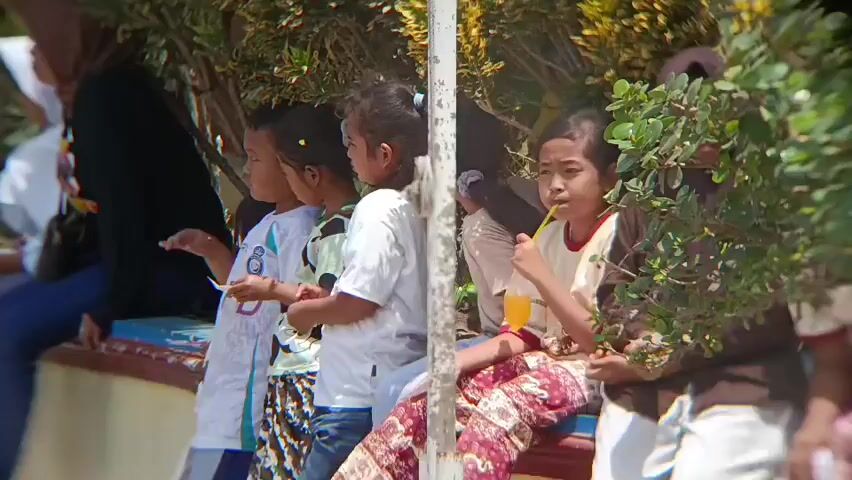 The video shows a group of children sitting on a bench outdoors. The children are of various ages and ethnicities. They are all dressed in casual clothing. Some of the children are holding drinks and snacks. The environment appears to be a park or a recreational area, with trees and plants in the background. The lighting suggests it is daytime, and the weather appears to be sunny. There are adults in the background, some of whom are also seated. The children seem to be interacting with each other and observing something off-camera.