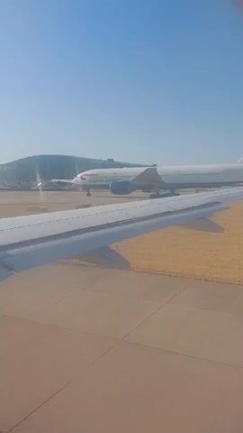 This video shows an aerial view of an airport tarmac on a clear, sunny day. In the foreground, the wing of an airplane is visible, extending from the bottom left to the middle of the frame. Beyond the wing, a second airplane, a British Airways Boeing 777, is parked on the tarmac. The airplane is predominantly white with blue and red livery. In the background, a modern airport terminal building with a glass facade and a curved roof is visible. Several other smaller vehicles and aircraft are also present on the tarmac. The overall scene depicts a typical airport environment with aircraft preparing for or having recently completed travel.