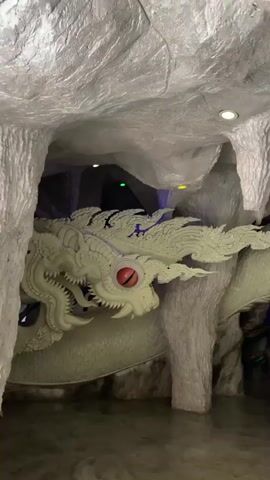 The video shows a large, pale green dragon sculpture inside a cave-like structure. The dragon has intricate scales and sharp, fin-like protrusions along its body and head. Its eye is a prominent red orb. The cave walls are textured and gray, resembling rock formations, with some lighting fixtures visible on the ceiling. A woman is partially visible on the right side of the frame, holding a phone up as if taking a picture or video. She is wearing a beige shirt and dark pants. The lighting in the cave is dim, with some spotlights illuminating the sculpture. The floor appears to be concrete. A fan is visible in the lower right corner of the frame.