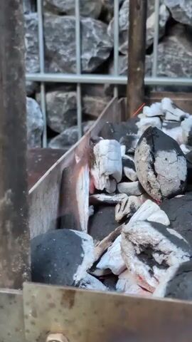 A close-up shot focuses on charcoal burning in a grill. The charcoal is grey and black, with some pieces glowing red. The grill appears to be made of metal. In the background, a stone wall is visible behind a metal fence. The scene is lit by natural light. There are no people or animals visible.