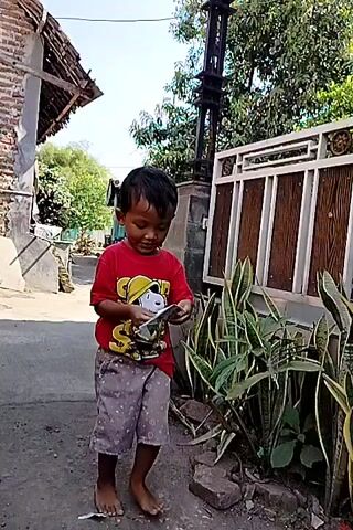 A young boy in a red t-shirt and patterned shorts stands outdoors, looking down at a white object in his hands. He is facing towards the right of the frame, and his body is partially obscured by a plant with long, green leaves. Behind him, a dark pole with visible electrical components rises vertically. To his left, a brick building with a corrugated roof is visible. To his right, a wooden fence with decorative elements runs horizontally. The ground appears to be a dirt or gravel path. The lighting suggests it is daytime, with shadows indicating sunlight.