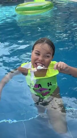 A young girl, wearing a bright neon green life vest over a swimsuit with pink floral designs, is in a swimming pool. She is holding a wooden skewer with several marshmallows on it and is in the process of eating one. The pool water is a clear blue, with ripples visible on the surface. In the background, a green and yellow inflatable raft floats on the water. The setting appears to be a sunny day, likely outdoors, given the bright water and raft. The girl's expression is one of mild enjoyment as she eats her treat. The visible text on her swimsuit reads "A N" in large pink letters. The scene takes place in Lastva Grbaljska, Montenegro.