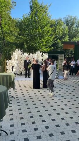 A wedding ceremony is taking place outdoors with a white flower archway in the background. A bride in a white dress and a groom in a suit stand in front of the arch. Several people are gathered around them, including a woman in a black dress who appears to be officiating. A photographer is filming the event. The ground is paved with black and white tiles. Trees and foliage are visible in the background, suggesting a garden setting. The weather appears to be sunny.