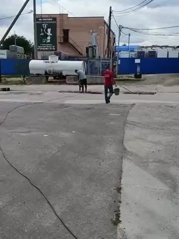 Two men are working on a damaged road. One man, wearing a grey shirt and blue shorts, is kneeling and appears to be using a tool on the road surface. The other man, wearing a red shirt and dark shorts, is standing and holding a bucket. They are near a white tanker truck, a transformer, and a construction fence. The scene is outdoors, with a cloudy sky.