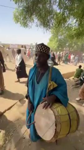 The video shows a man playing a drum in an outdoor setting. He wears a blue robe and a patterned cap. The drum has a yellow covering with dark stripes and is held by a strap across his shoulder. He uses two sticks to play the drum. In the background, there is a crowd of people dressed in various traditional garbs, walking on a dirt road. Some trees provide shade overhead, and the sky is clear.