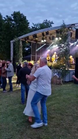 A group of people are gathered outdoors in a grassy area in front of a raised stage. The stage has a band performing with instruments and lights illuminating them. Some people in the foreground are dancing, while others are standing and watching the performance. The sky is overcast, suggesting it might be evening or a cloudy day. There are trees in the background.