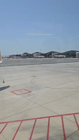 The video captures a bright, clear daytime scene at an airport in Abu Dhabi, United Arab Emirates, viewed from the window of a parked aircraft. Initially, the view shows a wide expanse of concrete tarmac with several airplanes parked in the distance, and the distinctive wavy roofs of modern airport terminal buildings under a clear blue sky.

As the camera pans right, the full wing and powerful engine of the viewer's aircraft come into view. The aircraft is an Etihad Airways plane, identifiable by its white fuselage adorned with large gold "ETIHAD" lettering and a distinctive patterned tail fin. A worker, dressed in a high-visibility vest, is seen on the tarmac near the aircraft's engine, performing ground operations or maintenance. A blue ground support unit and a white safety cone are positioned near the worker. The concrete tarmac features clear red-striped demarcation lines. Other aircraft are visible further away on the busy airport apron, underscoring the active environment of a major international airport.