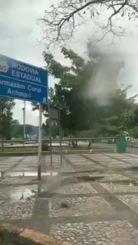 The camera is inside a vehicle, looking out at a park on a rainy day. The ground is wet and there are puddles on the pavement. Trees are visible and a motorbike drives by. There are bus stops and green fencing in the area.
