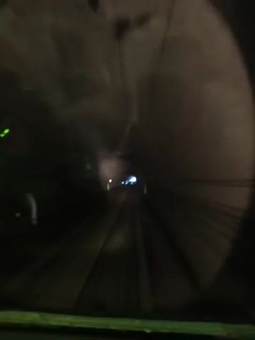 The video shows a point-of-view shot from inside a vehicle moving through a tunnel. The tunnel appears to be made of concrete and is dimly lit with lights visible in the distance. Tracks for the vehicle, likely a train, are visible on the ground. The view is framed by a circular opening, suggesting the shot is taken through a window or camera lens. The overall scene is dark and somewhat blurry, indicating movement. There are no people or other objects clearly discernible in the video, other than the tunnel structure and the tracks.