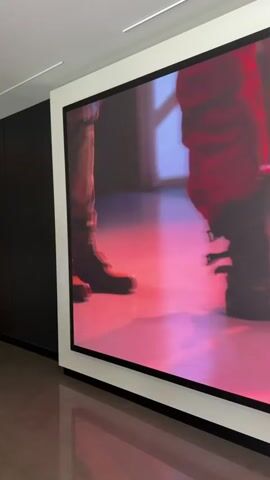 The video displays a series of large, framed images mounted on a wall. The first image shows the lower half of two people, one wearing a blue shoe and the other red, standing on a floor with a pinkish hue. The second image features a close-up of an astronaut's helmet and shoulder, with a person's hands adjusting it. The third image is an abstract shot of metallic tubes with a pink light reflecting off them. The fourth image is a blurry close-up of hands, possibly preparing to touch something. The final image is a close-up of a bright pink stylus, possibly a record player stylus, poised above a reflective surface, creating a pink reflection. The environment appears to be a gallery or exhibition space with a dark wall to the left and a white frame around the display.