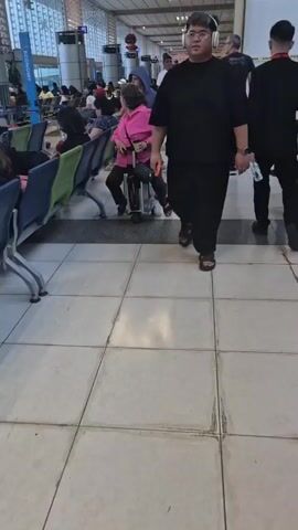 Inside an airport, passengers await their flights. A person walks past wearing a black outfit and sandals, large headphones around their neck. Another passenger sits on a wheeled suitcase, using it as a makeshift chair. Several other passengers are visible in the background, seated in waiting areas or walking through the terminal. The floor is tiled in a light color, and signage is visible overhead.