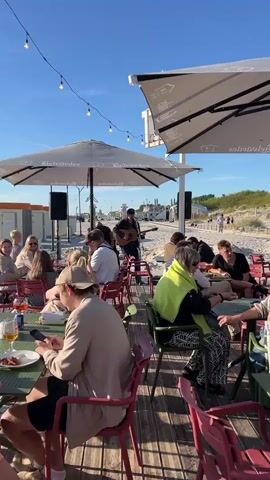 The video opens with a view of a beachside restaurant. Several people are seated at tables on a wooden deck. In the background, a musician is playing an acoustic guitar and singing into a microphone. Beyond the deck, there is a sandy beach with more people walking around and relaxing. The ocean is visible in the distance, with a clear blue sky overhead. As the video progresses, the camera pans to show more of the beach area, including some beach chairs and umbrellas. There are also some buildings in the background that appear to be beach facilities. The sun is shining brightly, casting long shadows. The overall atmosphere is relaxed and festive, with people enjoying the music and the beautiful beach setting.