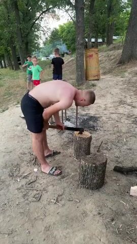 The video shows a man chopping wood outdoors. He is shirtless, wearing black shorts and flip-flops. He is standing over two tree stumps and swinging an axe into them. In the background, three children are visible. One child is wearing a green t-shirt, another a red t-shirt, and the third a black t-shirt. The environment appears to be a wooded area with a dirt path. A large metal container is visible on the right side of the frame. The sky is overcast. The overall atmosphere is casual, possibly a park or campground.