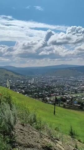 A drone or camera pans across a panoramic view of a town nestled in a valley. The town is characterized by a mix of houses with red and colorful roofs, interspersed with trees and green spaces. In the foreground, a grassy hillside slopes downwards, with a few scattered plants and some rocky outcrops. In the distance, rolling hills form the backdrop to the town, and the sky is a vibrant blue with scattered white clouds. The scene suggests a rural or semi-rural setting on a clear, sunny day.