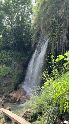 A waterfall cascades down a rocky cliff face overgrown with lush green vegetation. In the foreground, a wooden walkway crosses a shallow pool of water. A person in dark shorts stands near the edge of the pool on the left side of the frame. The surrounding environment is dense with trees and foliage, suggesting a natural, outdoor setting. The water from the waterfall creates a misty spray as it hits the rocks below.