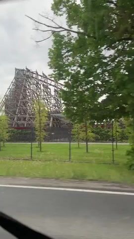 The video shows a drive-by view of a roller coaster from a car window. The roller coaster is visible behind trees and a fence, and appears to have both wooden and metal sections. The sky is overcast. The camera is moving, and at one point a blurred red car passes by. The overall scene is a glimpse of a roadside amusement park.