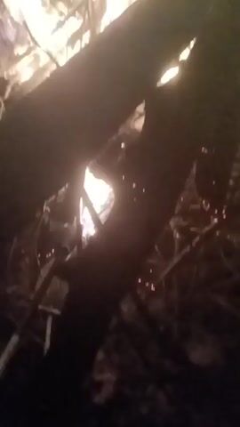 The video shows a close-up view of a bonfire at night. The fire is burning brightly, with flames reaching high into the air. Large pieces of wood are visible, supporting the fire. The surrounding area is dark, and the only source of light is the fire itself. The camera is positioned low, looking up at the fire. There is music playing in the background.