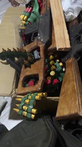 A partially clothed person sits in a chair holding a rifle, while a box containing an assortment of wooden toy weapons and ammunition is in the foreground on the floor. The setting seems to be indoors and cluttered.