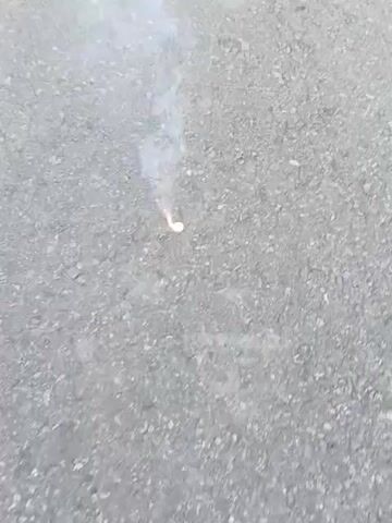 A firecracker sits on a gray asphalt surface and begins to burn, emitting white smoke and sparks. The surface is uneven, with small pebbles visible. A small, white object is visible next to the firecracker before it ignites. The firecracker burns intensely, producing a cloud of smoke around it.