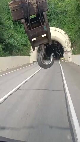 The video captures a POV perspective of driving through a tunnel. The primary object of interest is a large, metallic hook hanging from what appears to be a crane or industrial equipment, positioned prominently in the foreground, slightly to the left. The hook is substantial, with a weathered, metallic texture.

The scene is a two-lane road, marked with white lines, leading into a tunnel. The tunnel entrance is brightly lit with an arched ceiling, and as the vehicle progresses, the tunnel interior becomes darker, illuminated by a series of lights along the ceiling. The walls of the tunnel appear to be concrete or rock. Vegetation, specifically green trees and foliage, is visible on the hillsides surrounding the tunnel entrance, suggesting a natural, possibly mountainous environment.

Several cars are visible within the tunnel, indicating ongoing traffic. One silver SUV is particularly noticeable as it passes by on the left lane. The lighting conditions suggest it is daytime, but the exact time is not discernible. The weather appears to be clear, based on the visibility and the lush greenery.

There are a few signs visible near the tunnel entrance, including a circular sign with a red border and a diagonal line, which typically signifies a prohibition. There are also some yellow and black striped markers, common for road hazards. The overall mood is one of transit and perhaps a touch of industrial presence due to the large hook.

The specific location is identified as Kazachiy Brod, Russia. There is no discernible text within the video itself, apart from the prohibition signs.