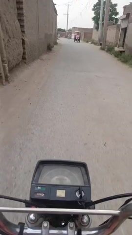 The video shows a first-person perspective from a person riding a motorcycle down a narrow, paved road in what appears to be a rural area. The road has some wear and patches. On either side of the road are buildings, some brick or concrete, and sparse vegetation. In the distance, other vehicles, including a motorcycle and a small three-wheeled vehicle, are visible. The sky is overcast. The motorcycle's handlebars and instrument panel are visible in the foreground.