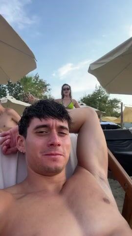 A man with dark hair and bare chest reclines in a lounge chair on a sandy beach, looking at the camera. Behind him, a woman in a lime green bikini stands with her arms outstretched, facing the camera. She wears sunglasses and jewelry. Another person is partially visible to the left. Beach umbrellas and lounge chairs are scattered in the background. The sky is clear and blue. The woman leans down towards the man, her face animated.