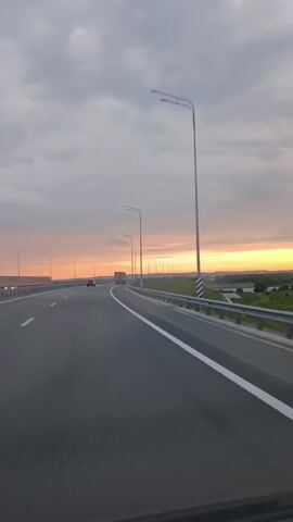 The video shows a view from inside a moving vehicle on a highway at sunset. The sky is filled with clouds tinged with orange and yellow from the setting sun. A highway with two lanes is visible, with a guardrail separating the lanes from the right side. Streetlights are spaced along the highway. In the distance, there are fields and trees. A red car and a truck are visible on the highway ahead. The motion blur indicates the vehicle is moving at a moderate speed.